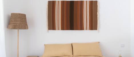 A bedroom with a bed, a wall-mounted striped tapestry, a floor lamp, and a bedside table with a lamp.