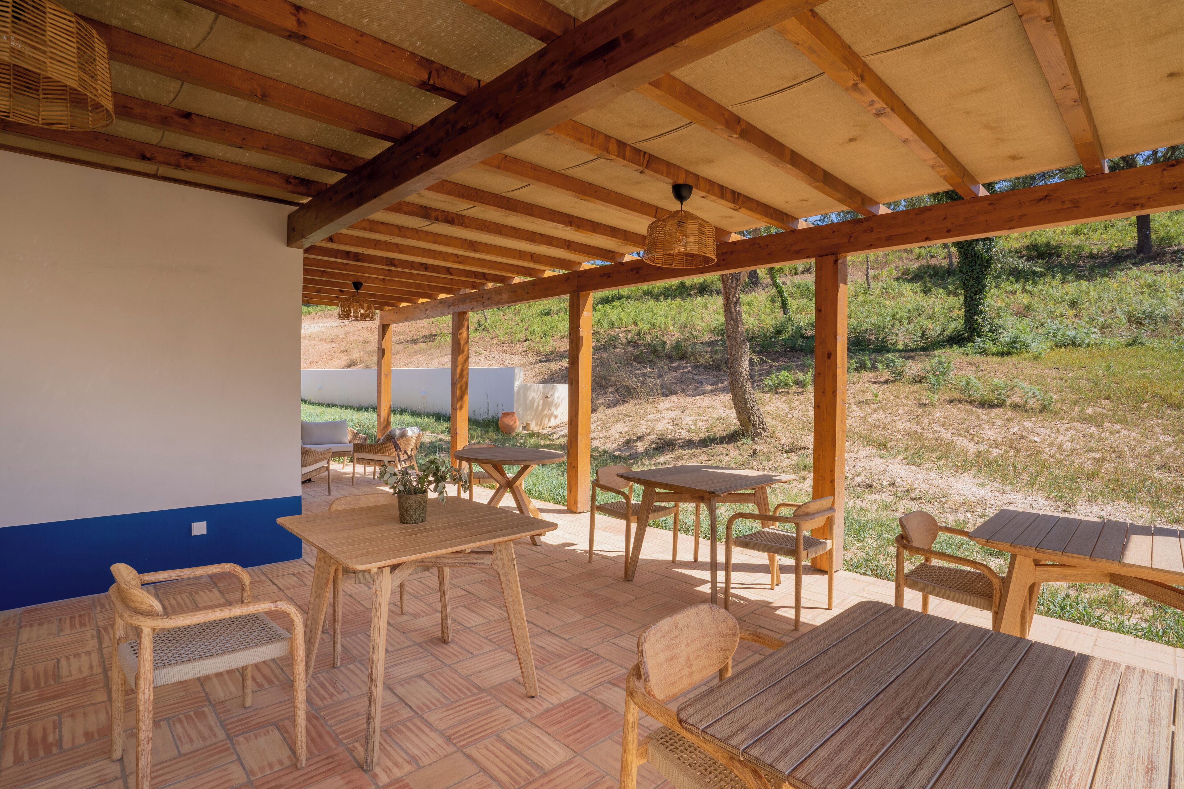An outdoor patio with wooden tables and chairs, a covered area with a wooden ceiling, and a view of a grassy landscape.