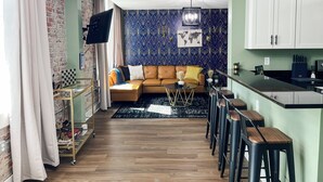 Una sala moderna con un sofá de cuero marrón, una mesa de centro negra y un muro estampado. Hay una zona de cocina con armarios blancos y un taburete de bar.