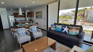 A modern living room with a sofa, armchairs, and a coffee table. There is a kitchen area with cabinets and a dining table. Large windows offer a view of buildings and greenery outside.