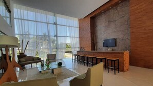 A modern living room with a glass coffee table, sofas, a TV mounted on a stone wall, and a bar area with stools. - Peninsula  Charming and luxury apartment on the beach, with beautiful ocean view (Mazatlán)