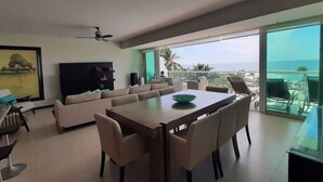 A modern living room with a large sofa, dining table, and a TV. There is a balcony with a view of the ocean. - Peninsula  Charming and luxury apartment on the beach, with beautiful ocean view (Mazatlán)