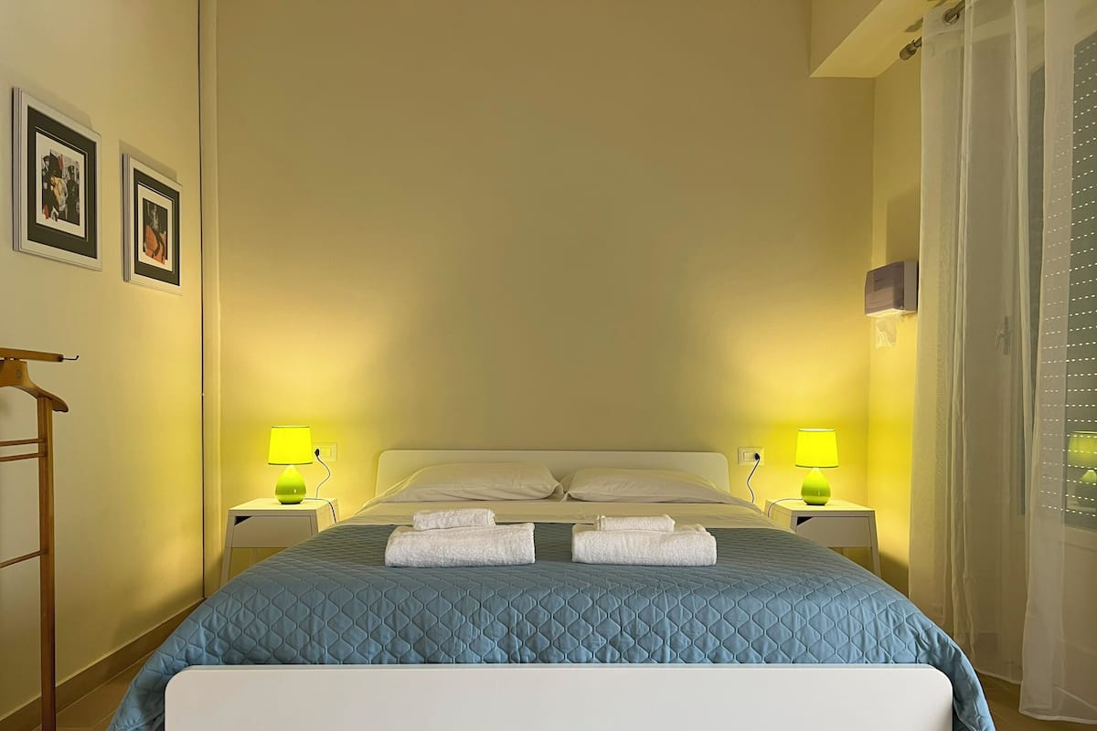 Una cama bien hecha con un edredón azul, dos lámparas de noche y un perchero de madera a la izquierda.