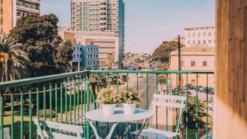 A balcony with a table and chairs, overlooking a city street with buildings and parked cars.
