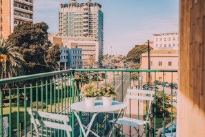 Un balcon avec une table et des chaises, donnant sur une rue de la ville, avec des immeubles et des voitures garées.