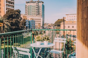 Un balcón con mesa y sillas, con vistas a una calle urbana con edificios y coches estacionados.