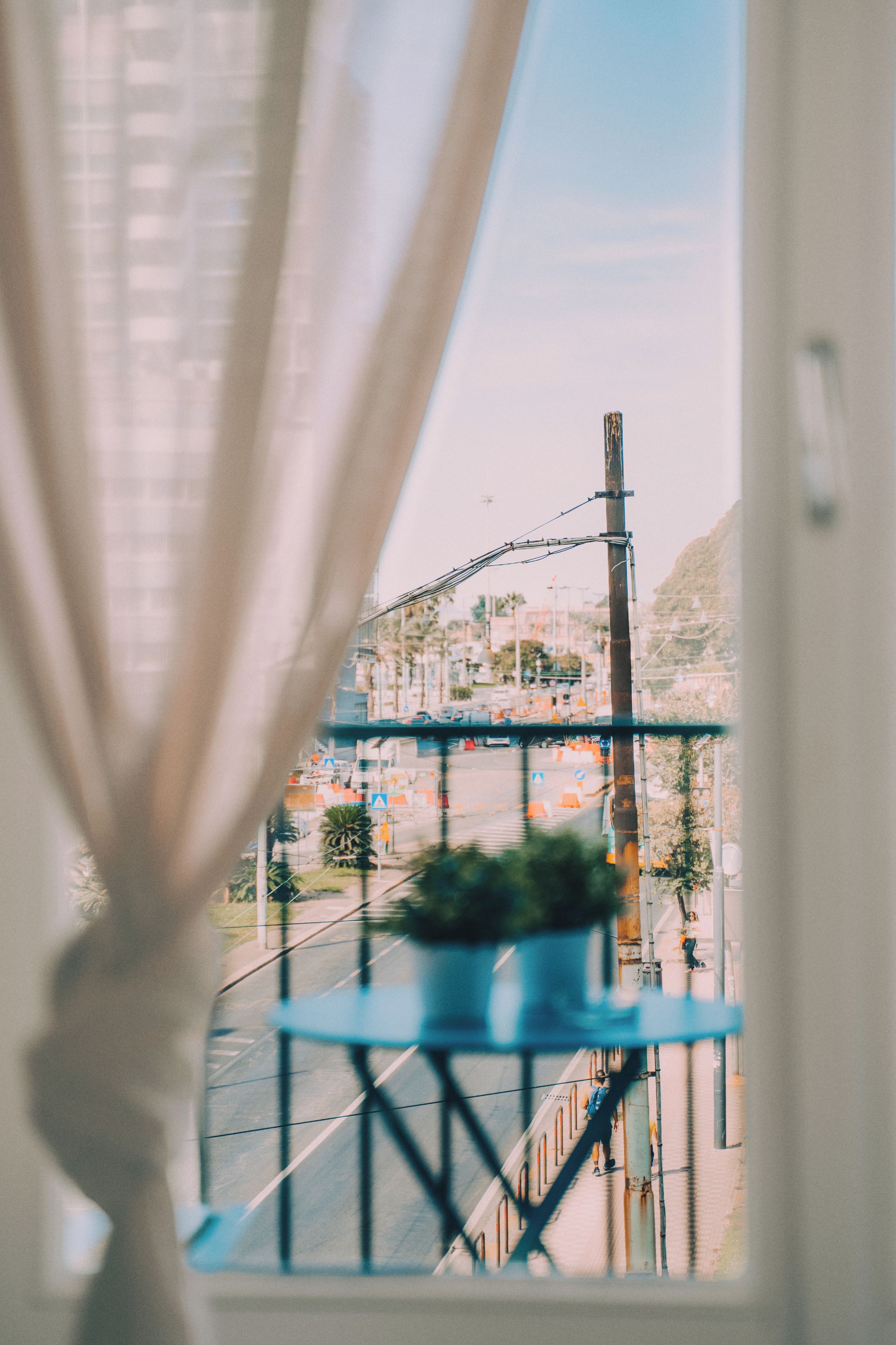 A view through a window with a curtain, featuring a balcony with a potted plant, a street with traffic, and a mountain in the background.