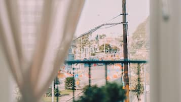 A view through a window with a curtain, featuring a balcony with a potted plant, a street with traffic, and a mountain in the background.