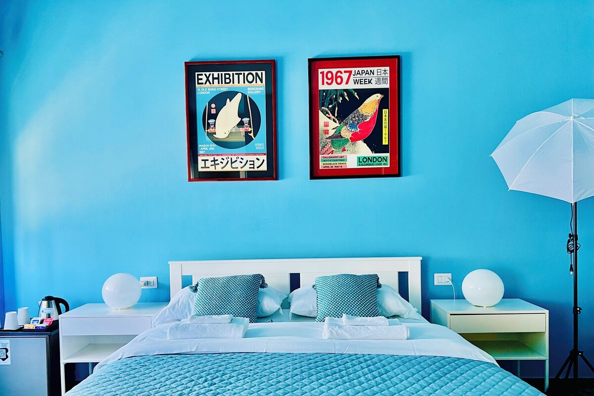 A hotel room with a blue wall, a bed with a blue quilted bedspread, two framed posters, a white lamp, and a white umbrella.