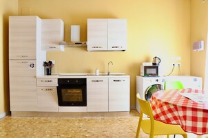 Cocina compacta con microondas, horno y lavabo. Hay una mesa con mantel a cuadros y una silla.