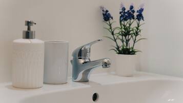 Un lavamanos con grifo cromado, dispensador de jabón blanco y taza blanca. Se ve una planta en maceta con flores azules al fondo.