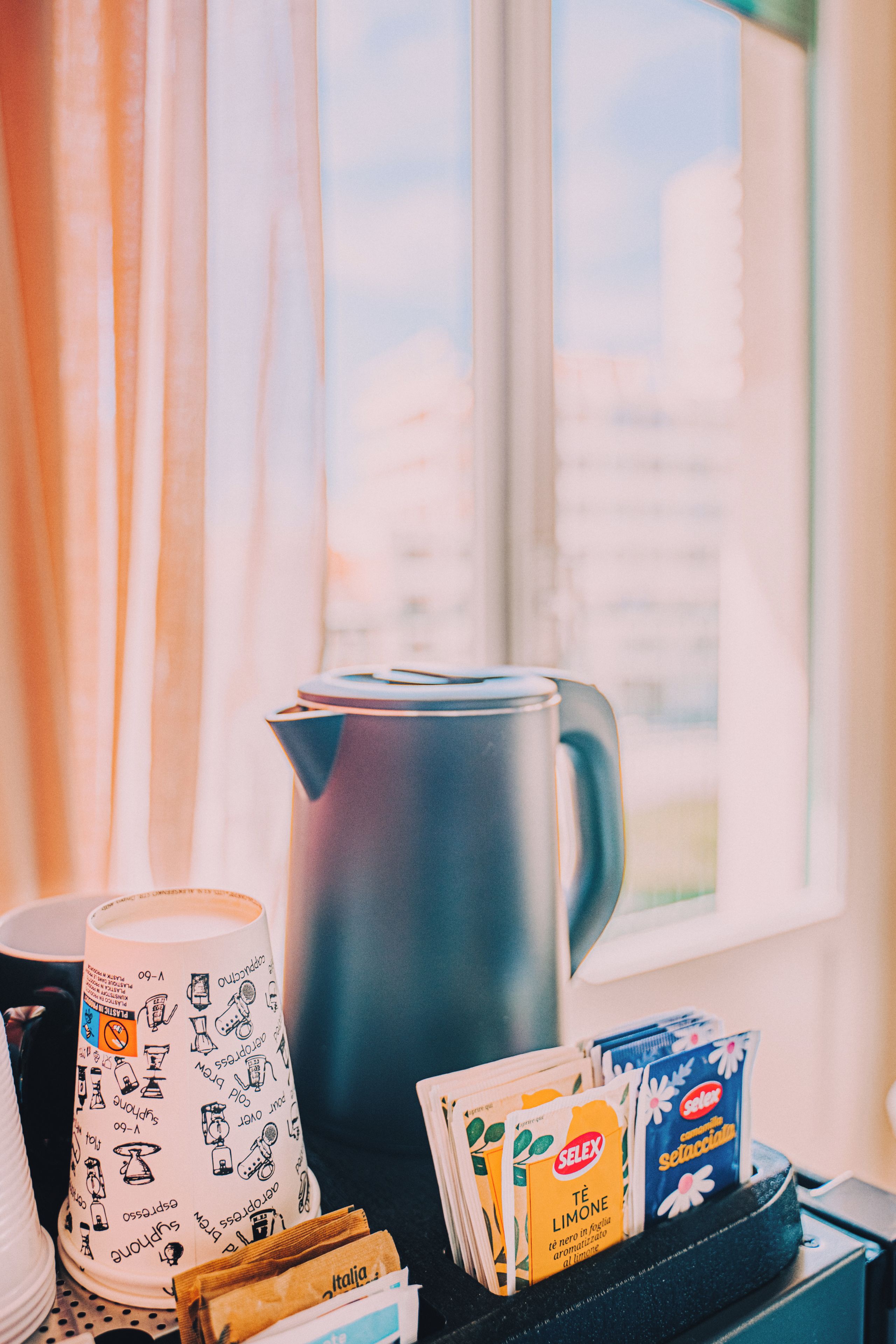 Une machine à café, une tasse et des sachets de thé sont posés sur une table près d’une fenêtre ornée de rideaux.
