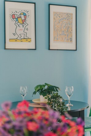 Una habitación con dos cuadros enmarcados en la pared, una pequeña mesa redonda con una planta en maceta y dos copas de vino, y vista a una pared azul.