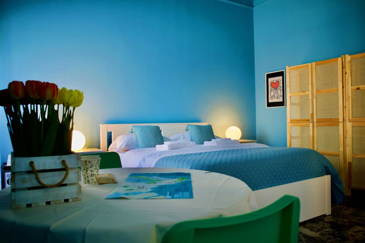Un dormitorio con cama, mesita de noche, un jarrón con flores y un cuadro en la pared.