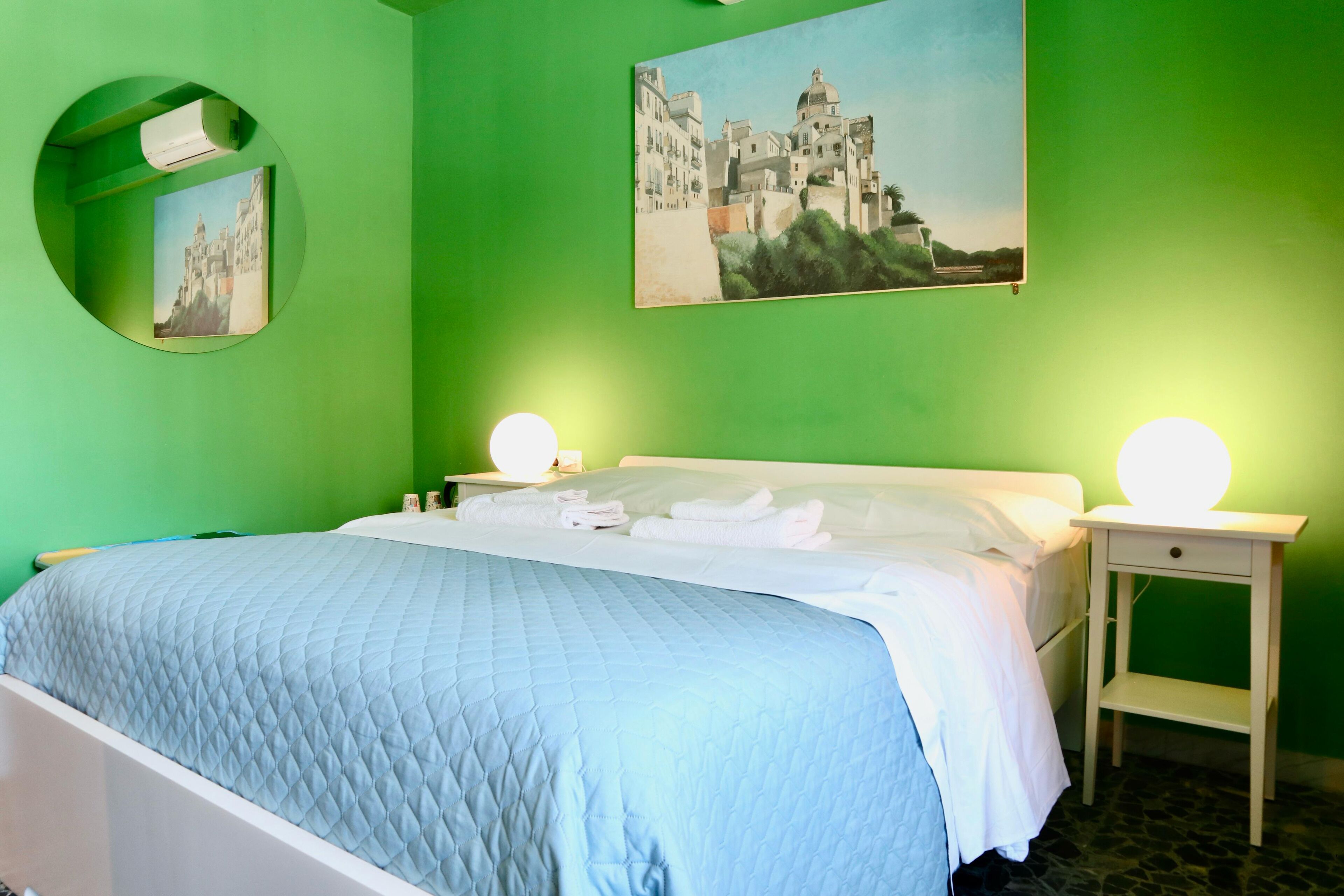 A neatly made bed with a blue quilt, two bedside tables with round lamps, a green wall, and a painting of a building.