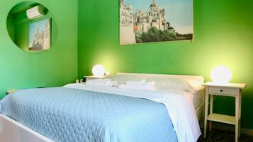 A neatly made bed with a blue quilt, two bedside tables with round lamps, a green wall, and a painting of a building.