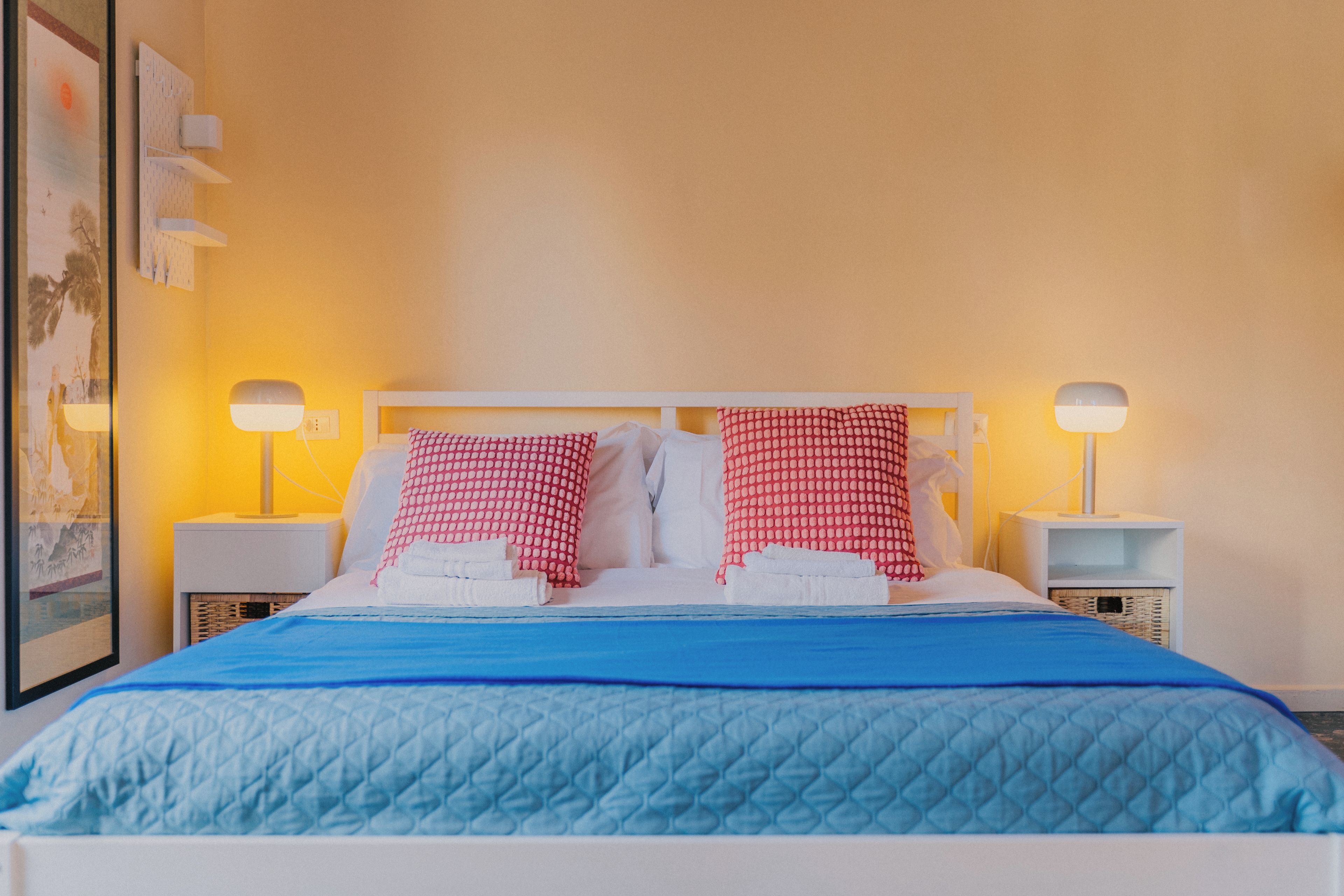 A hotel room with a bed, two bedside lamps, a framed picture on the wall, and a blue bedspread.