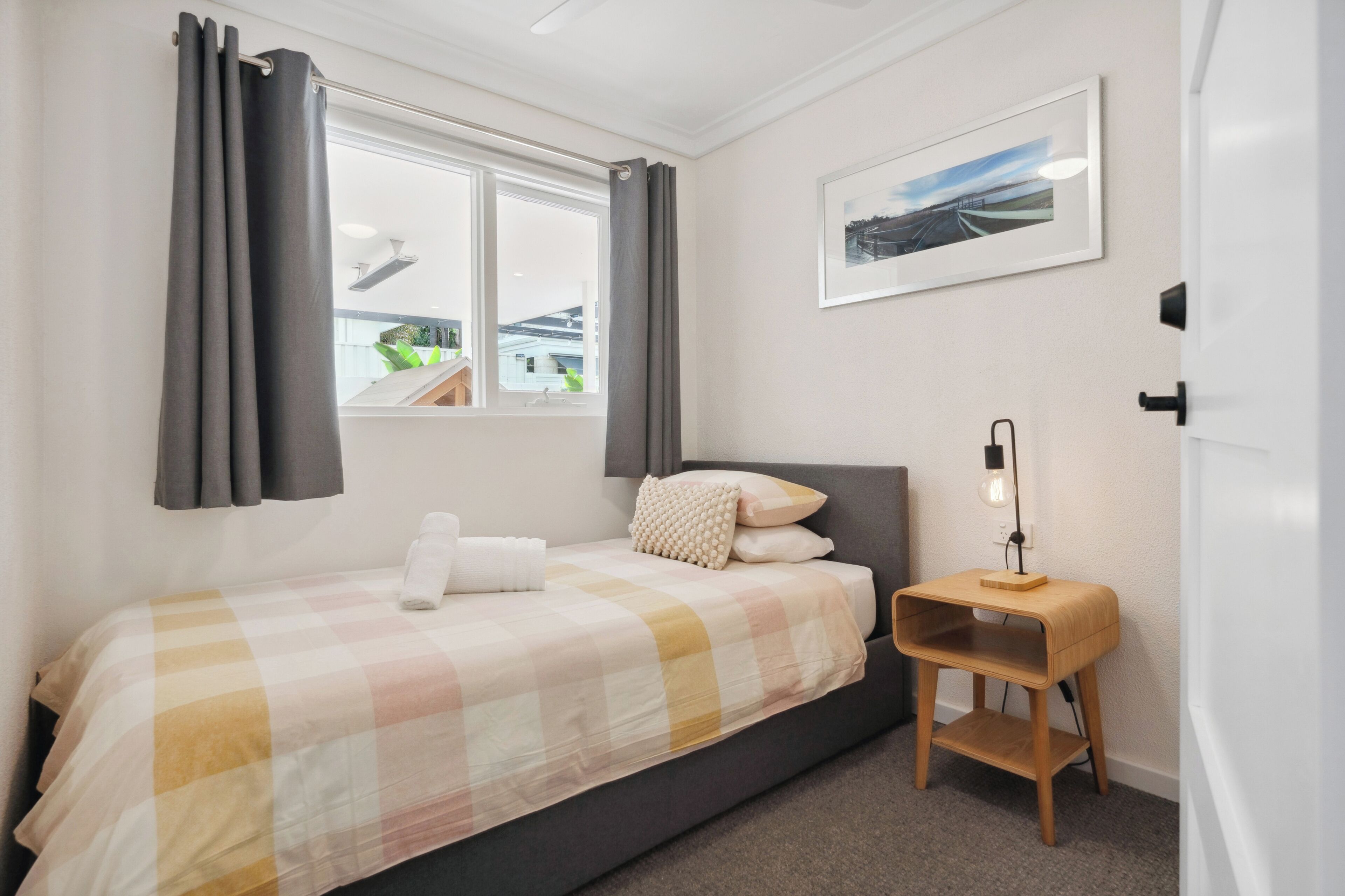 Un dormitorio con cama, cortinas, una mesita de noche con lámpara y un cuadro en la pared.
