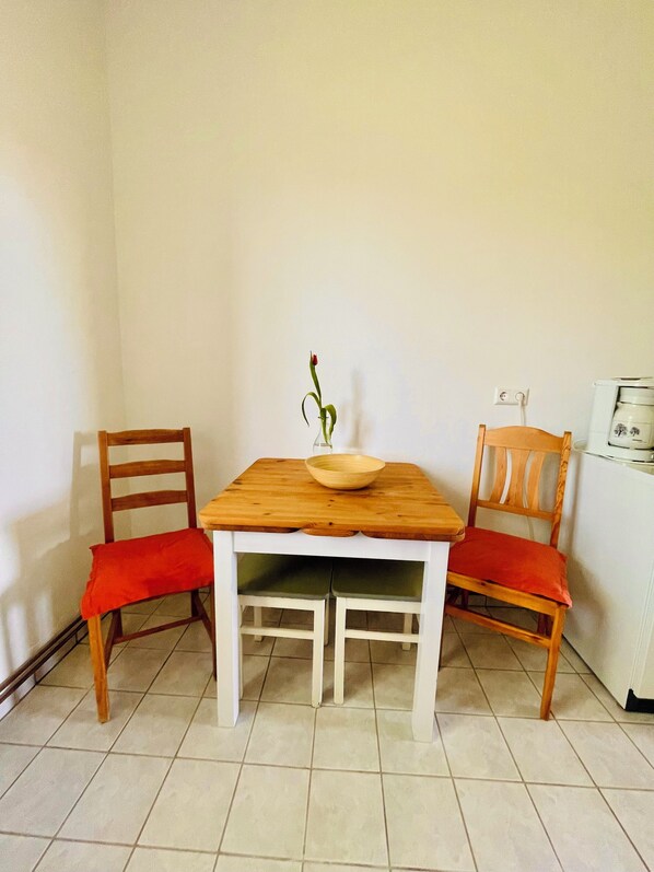 Una mesa de comedor con tapa de madera y patas blancas, flanqueada por dos sillas de madera con cojines rojos. Un jarrón con una planta está centrado en la mesa.