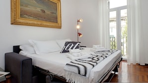 Cama bem arrumada com lençóis brancos, manta preta e branca e almofada com design marcante. Um quadro está pendurado acima da cama, e um abajur com três lâmpadas está posicionado ao lado.