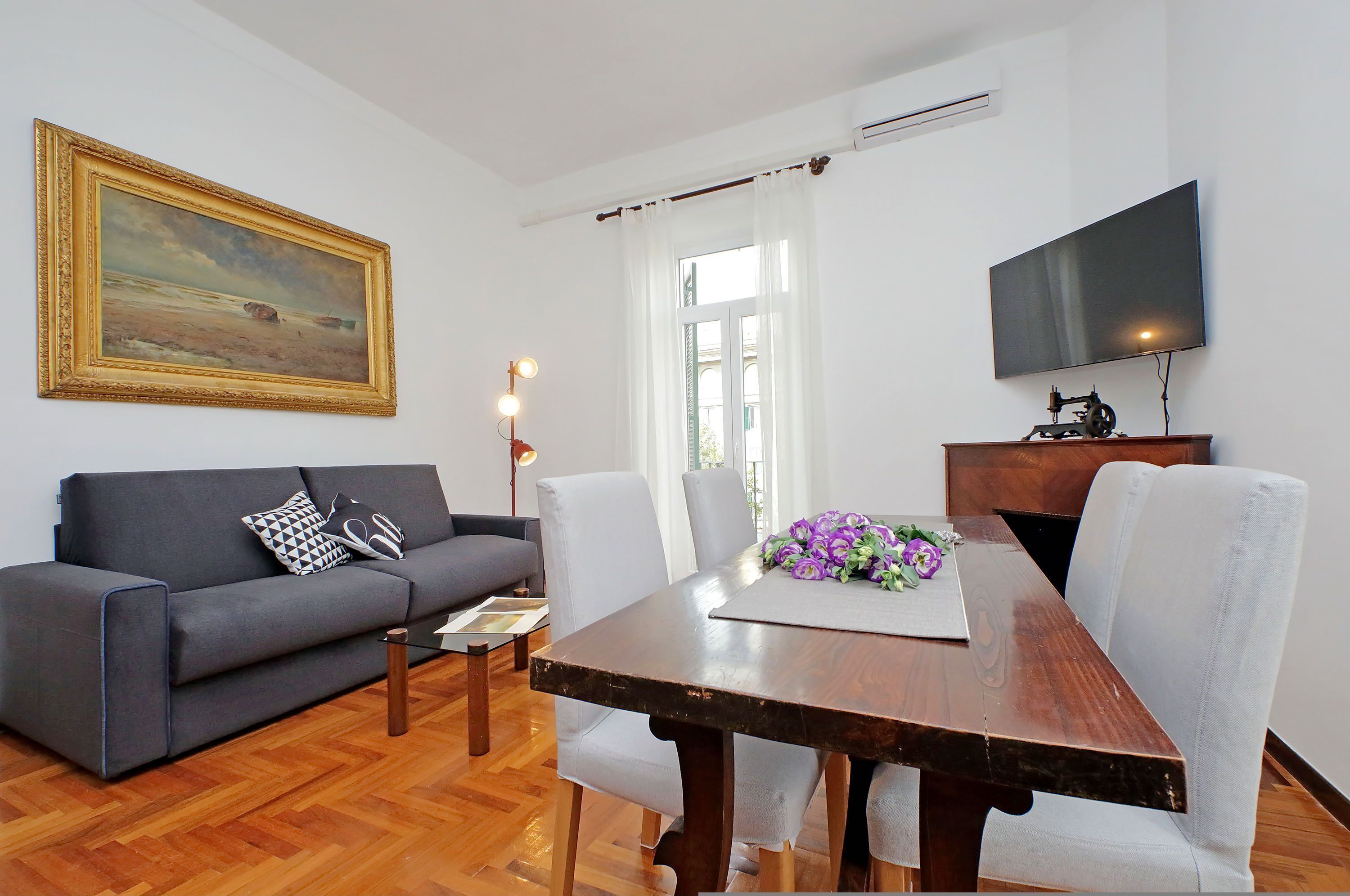 A living room with a sofa, dining table, chairs, a painting, and a TV.