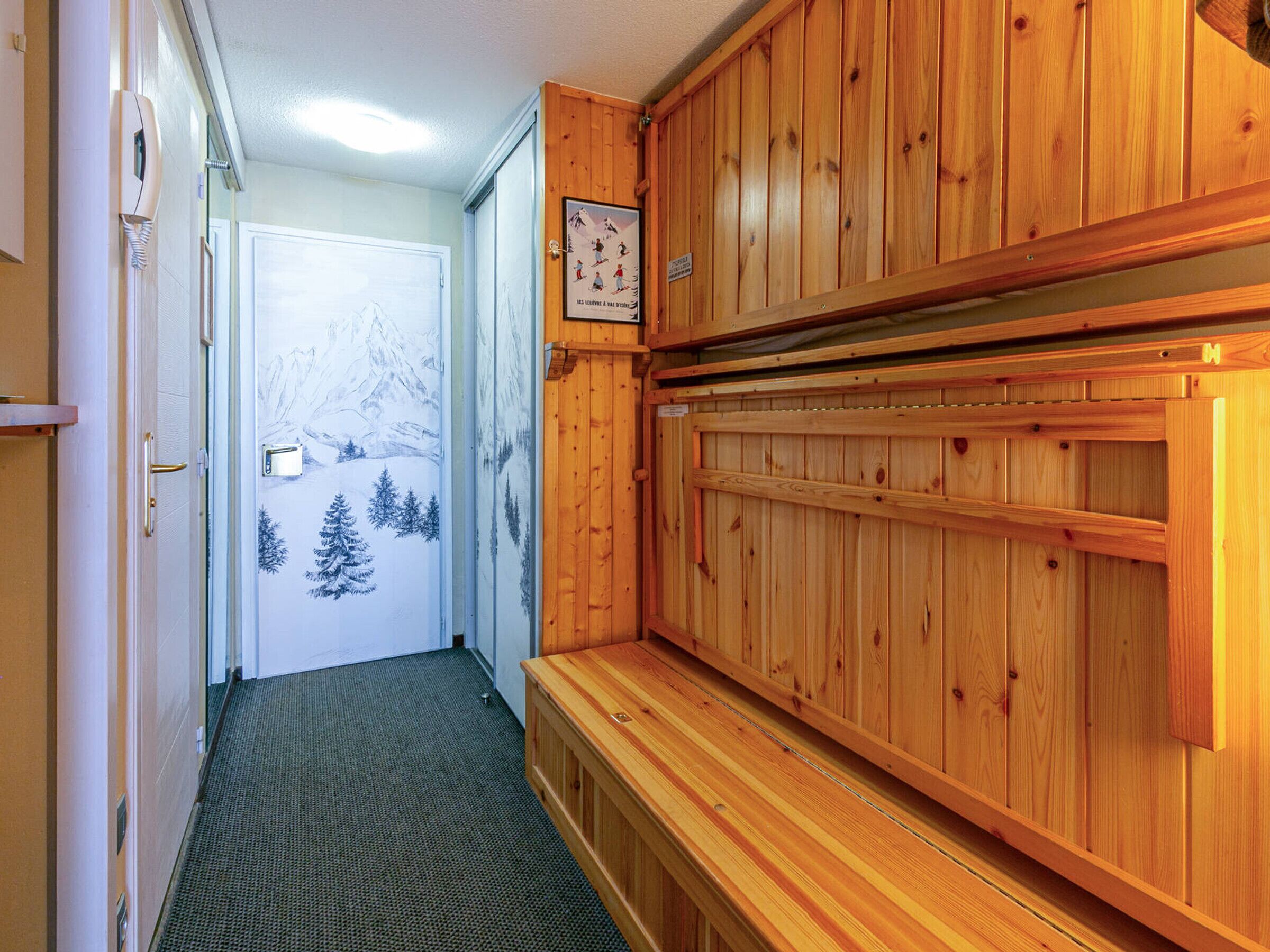 A wooden cabin interior with a door leading to a snowy outdoor scene, a framed picture on the wall, and a bench.