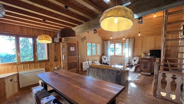 A rustic wooden interior with a large wooden dining table, benches, and a bar area. There are multiple windows providing natural light, and a ladder is visible in the background.