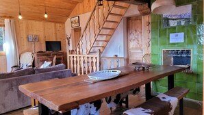 A rustic wooden interior with a wooden staircase, a dining table with a cowhide bench, a sofa, and a television in the background.