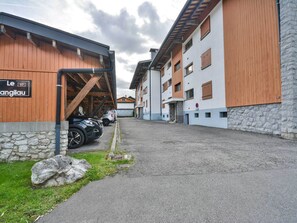 A building with a sign reading 'Le 97 angilau,' a parking area with cars, and a stone wall.