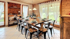 A dining area with a wooden table set for six, surrounded by chairs, with a wooden ceiling and large windows with curtains. - Cozy 4* chalet near slopes with parking and wifi for 14 guests (Le Corbier)
