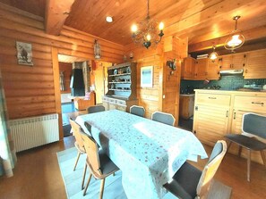 Une cabane en bois comprenant un coin repas avec une table et des chaises, une cuisine avec des meubles en bois et un lustre.