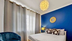 Un dormitorio con cama, un sillón azul, una mesita de noche con lámpara y un adorno en la pared.