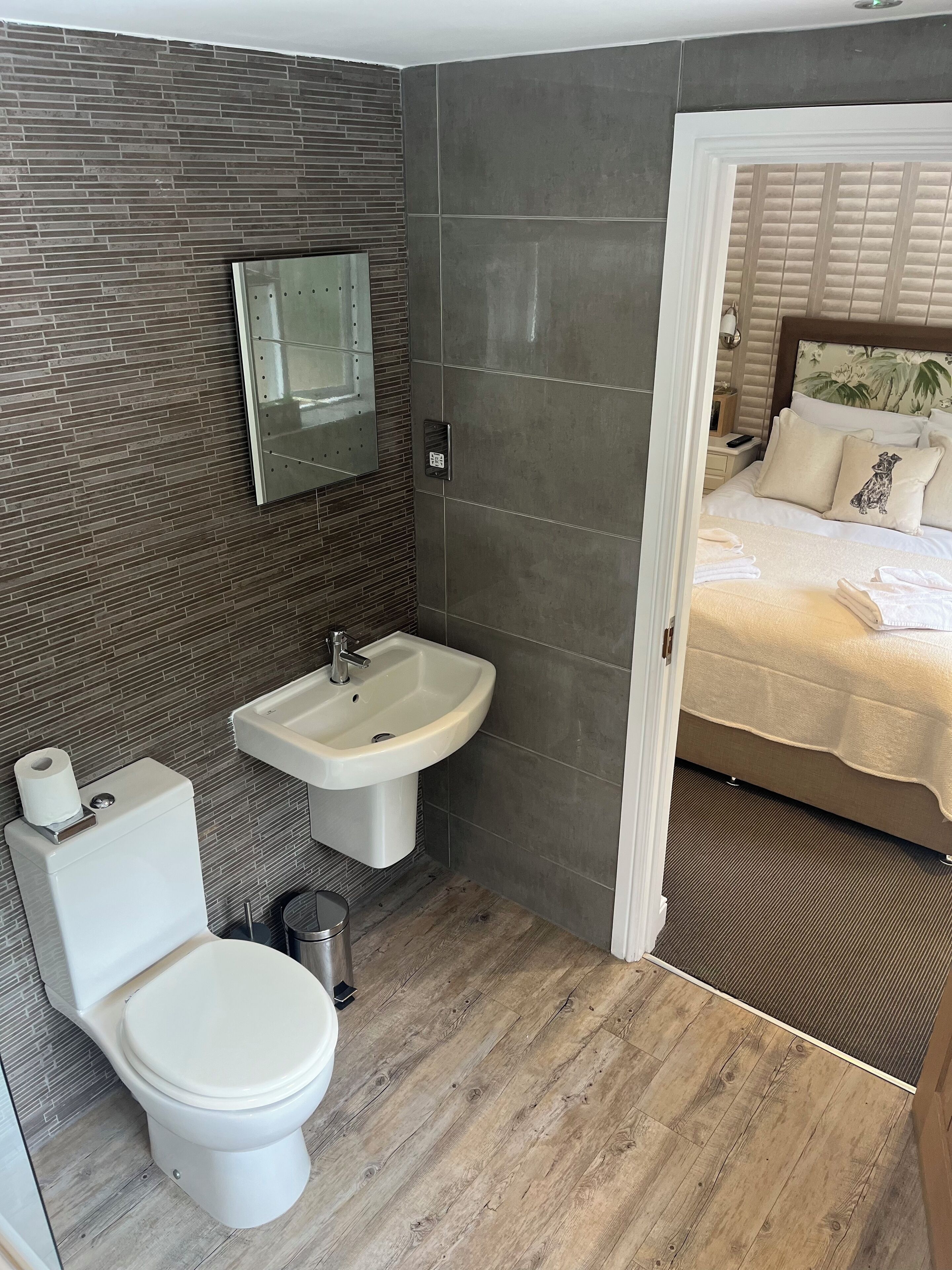 A compact bathroom with a toilet, sink, and mirror. A bed with a headboard and pillows is visible through an open door.