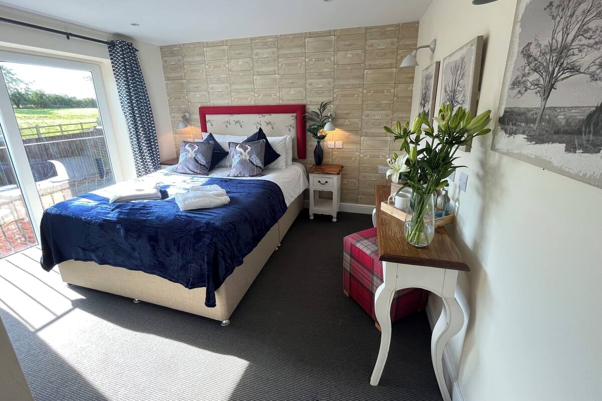 A bedroom with a large bed, a bedside table, a nightstand, a window with a view, and a wall with artwork.