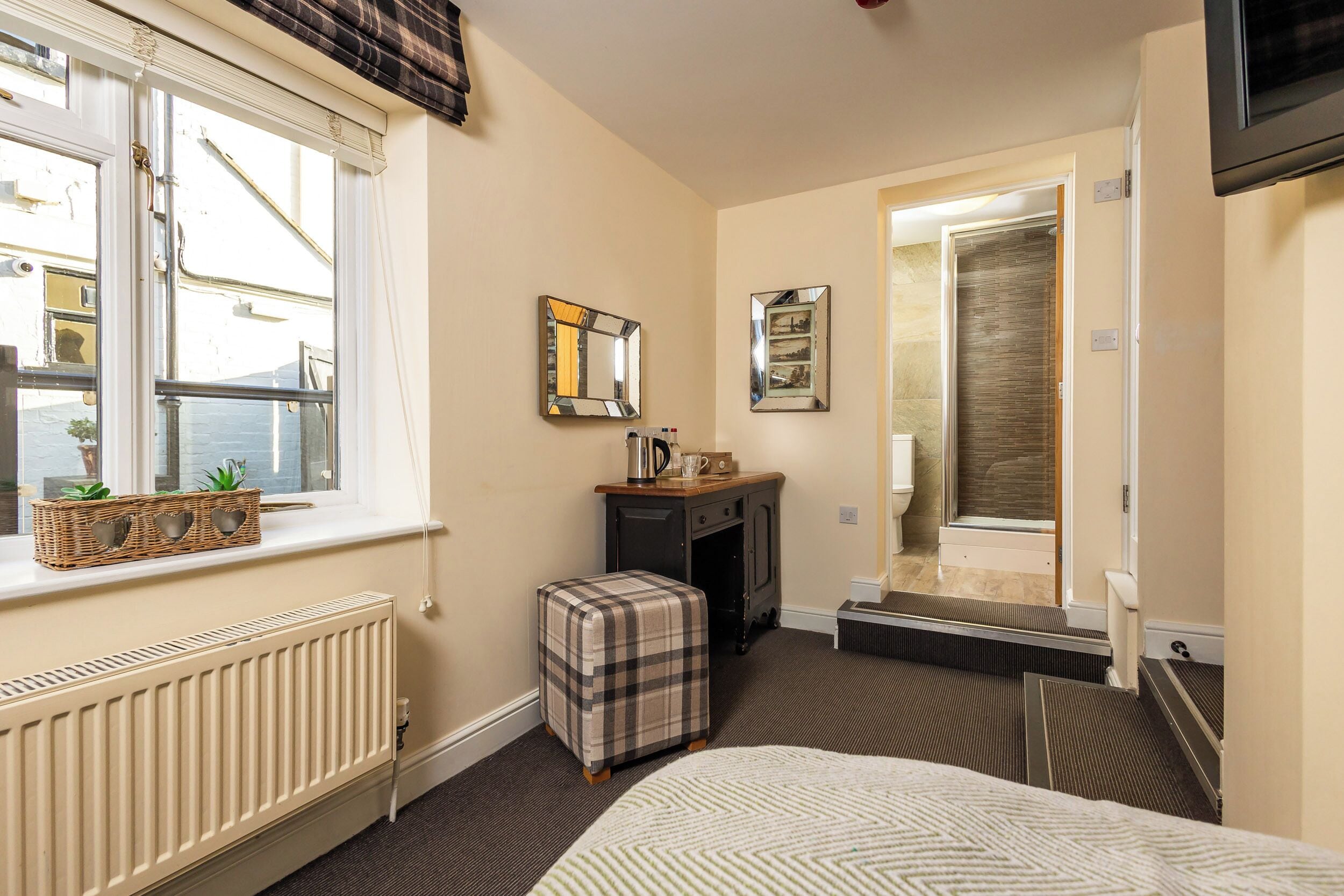 A compact hotel room with a bed, a small desk with a mirror, a radiator, a window with a view, and a door leading to a shower area.
