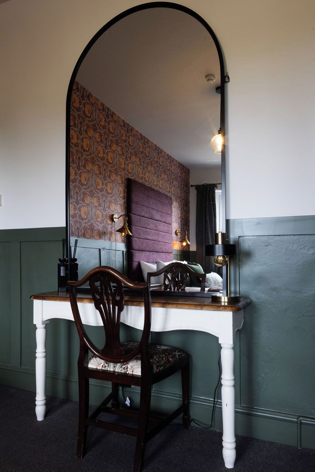 A room with a desk, a chair, a mirror, and a patterned wallpaper.