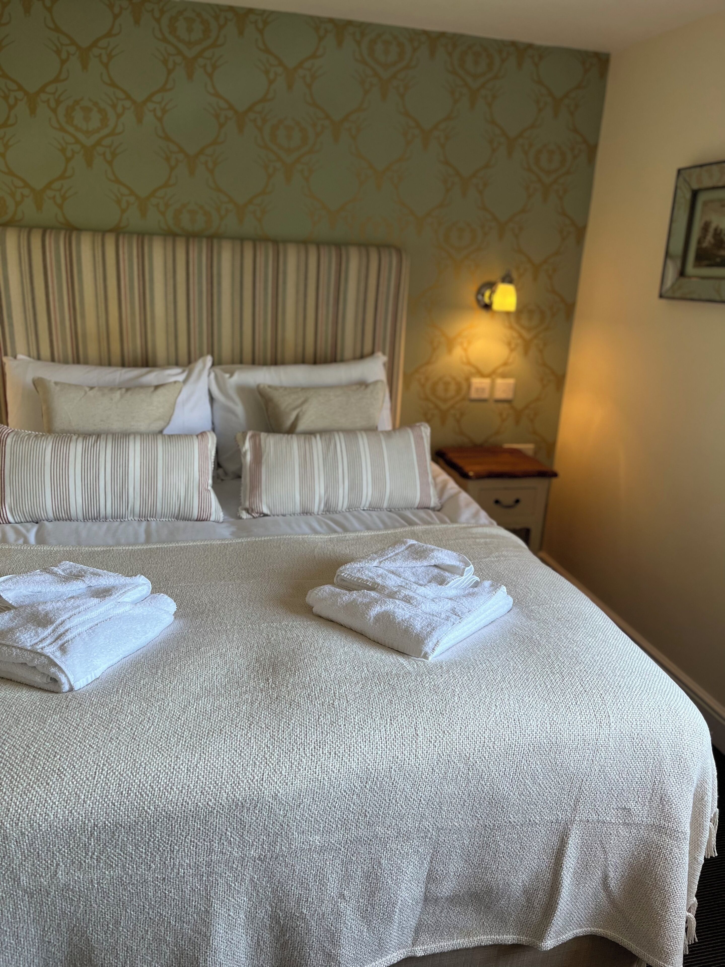 Een net opgemaakt bed met witte lakens en kussens, een bedlampje en een behang met een patroon.