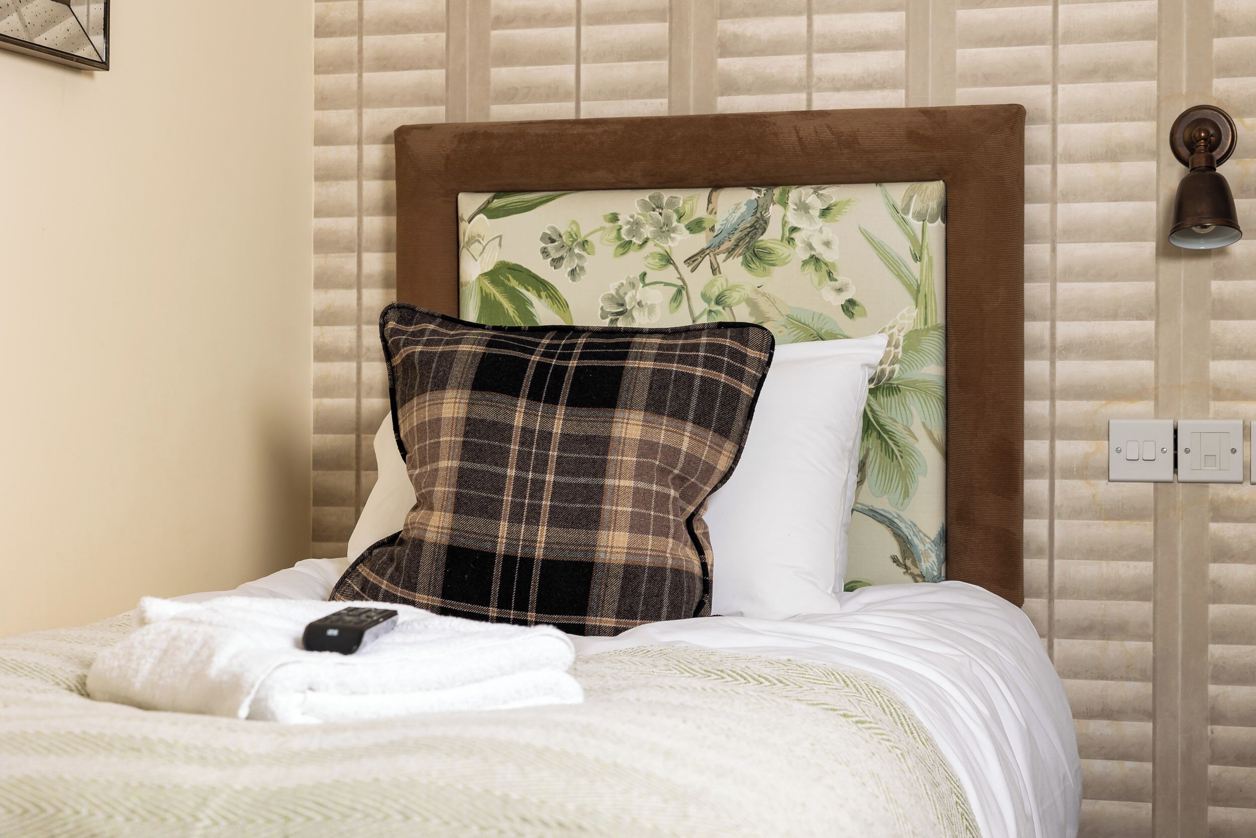 A neatly made bed with a remote control, a plaid pillow, and a floral headboard.