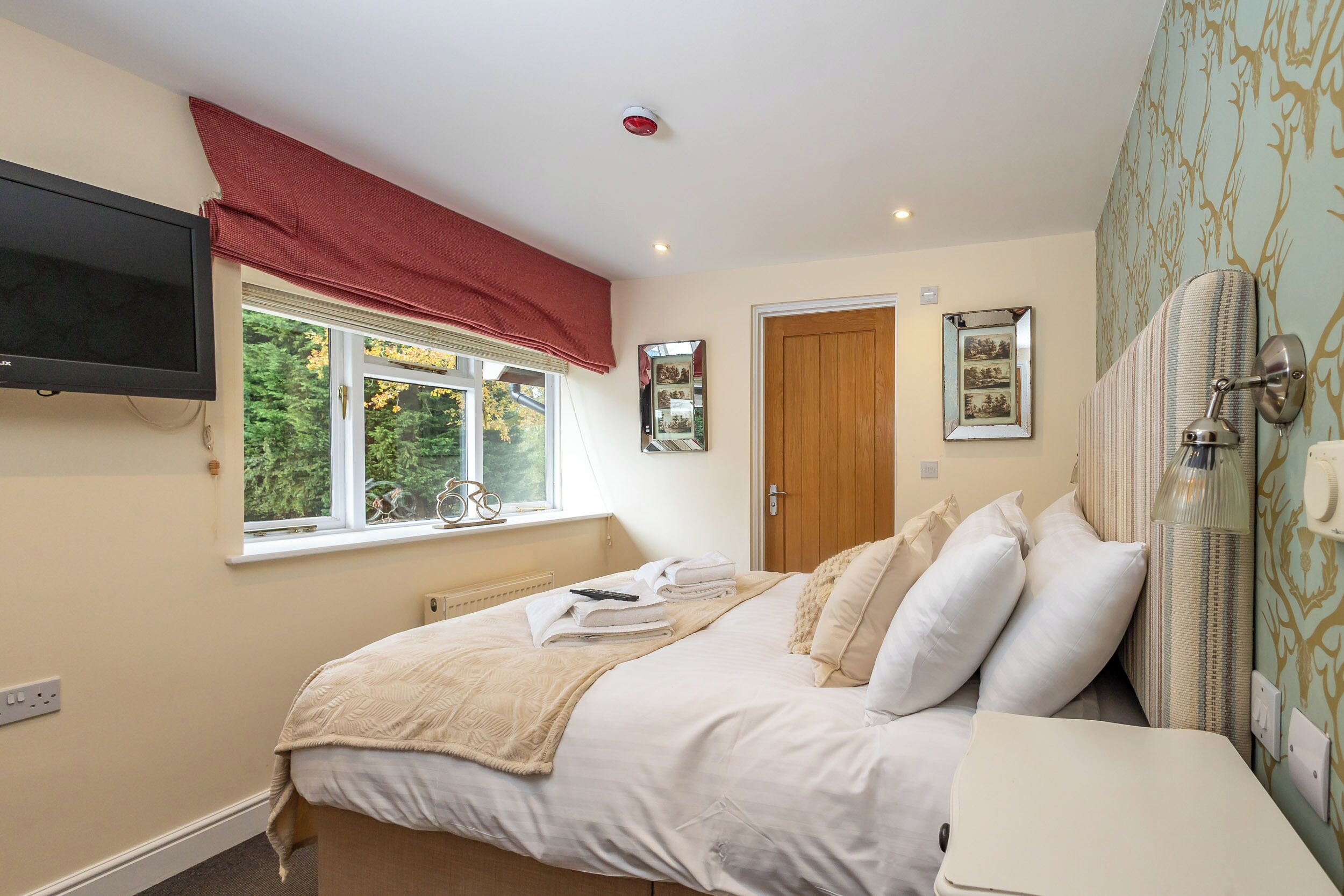 A bedroom with a bed, a TV mounted on the wall, a window with a red valance, and a door.