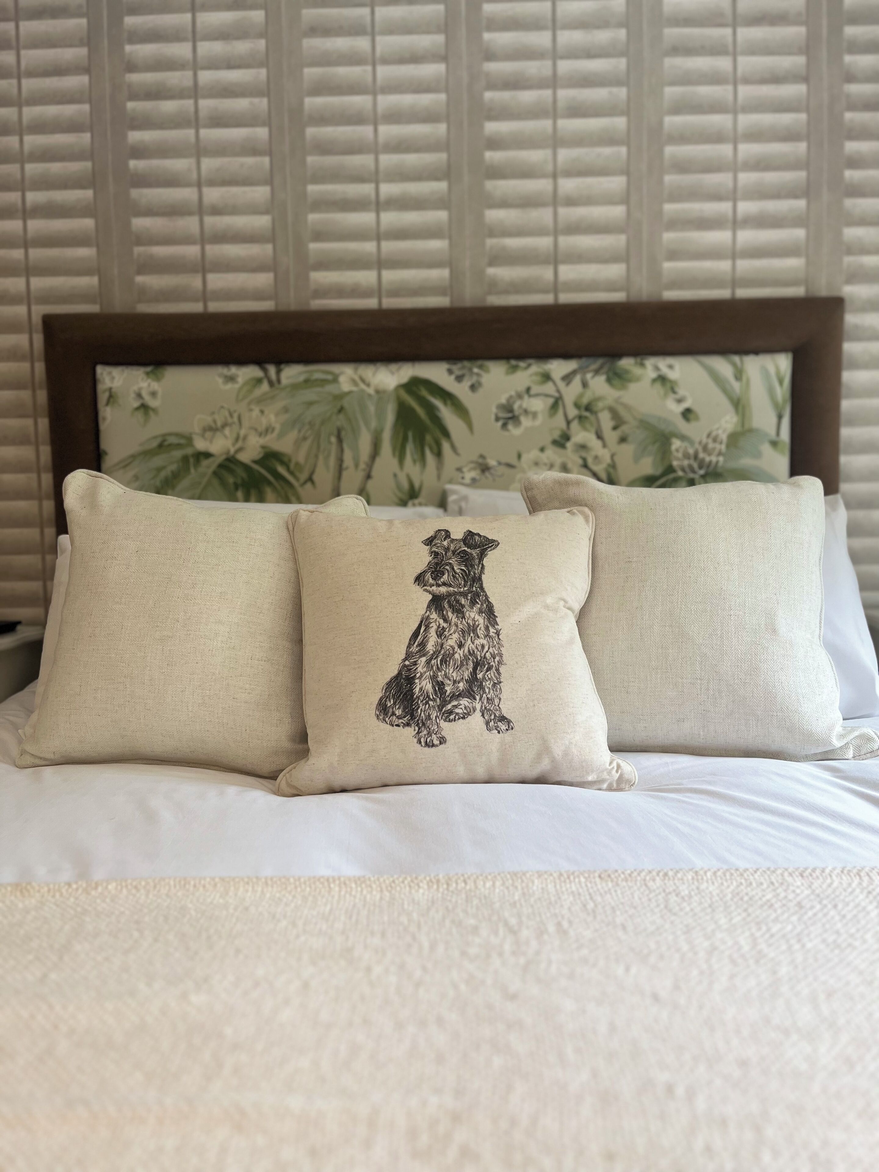 Una cama con tres almohadas, una con una ilustración de un perro, frente a una pared con papel tapiz floral y una ventana con persianas.