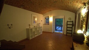 Une pièce spacieuse avec un plafond en briques, des murs blancs et un sol carrelé. On y trouve un banc en bois, une étagère blanche et une porte donnant sur l’extérieur.