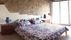 Une chambre avec un mur en pierre, un lit recouvert d’une couvre-lit à motifs floraux, deux tables de chevet et une fenêtre.