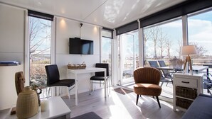Una stanza moderna e luminosa con una grande finestra, una TV a schermo piatto, un'area pranzo e un salotto con una lampada.