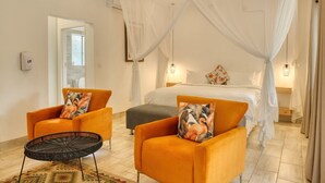 Une chambre à coucher avec un lit, deux fauteuils orange, une petite table ronde et un lit à baldaquin avec des rideaux blancs.