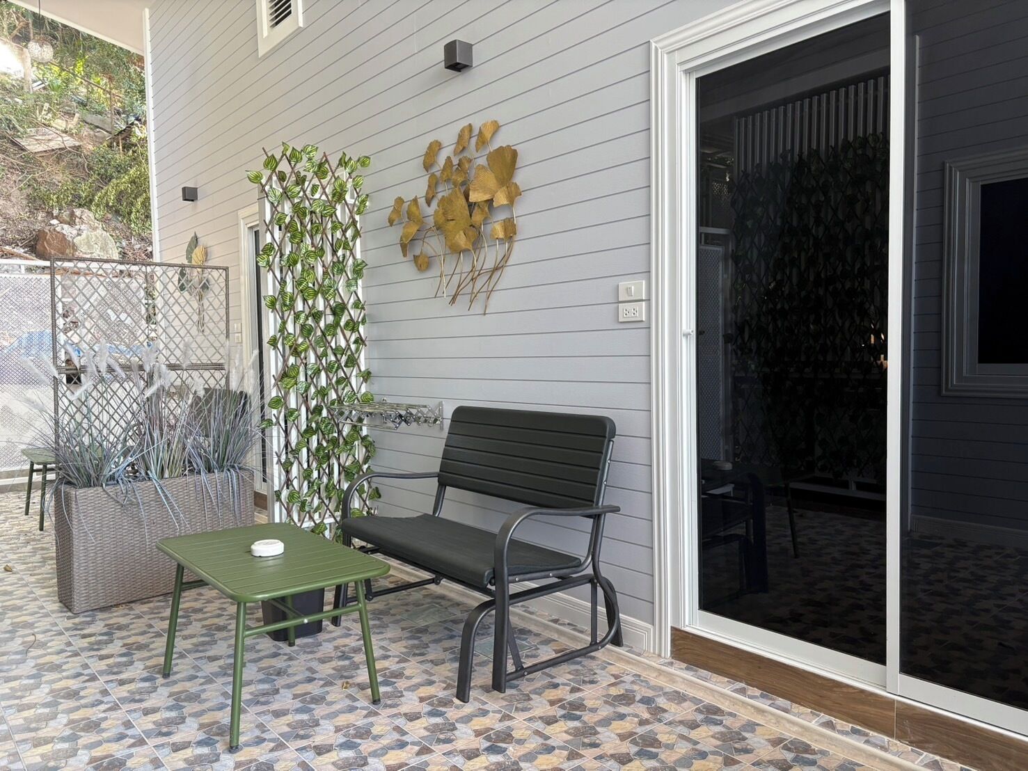A patio with a green table and a black bench, a decorative wall with leaf patterns, and a sliding glass door.