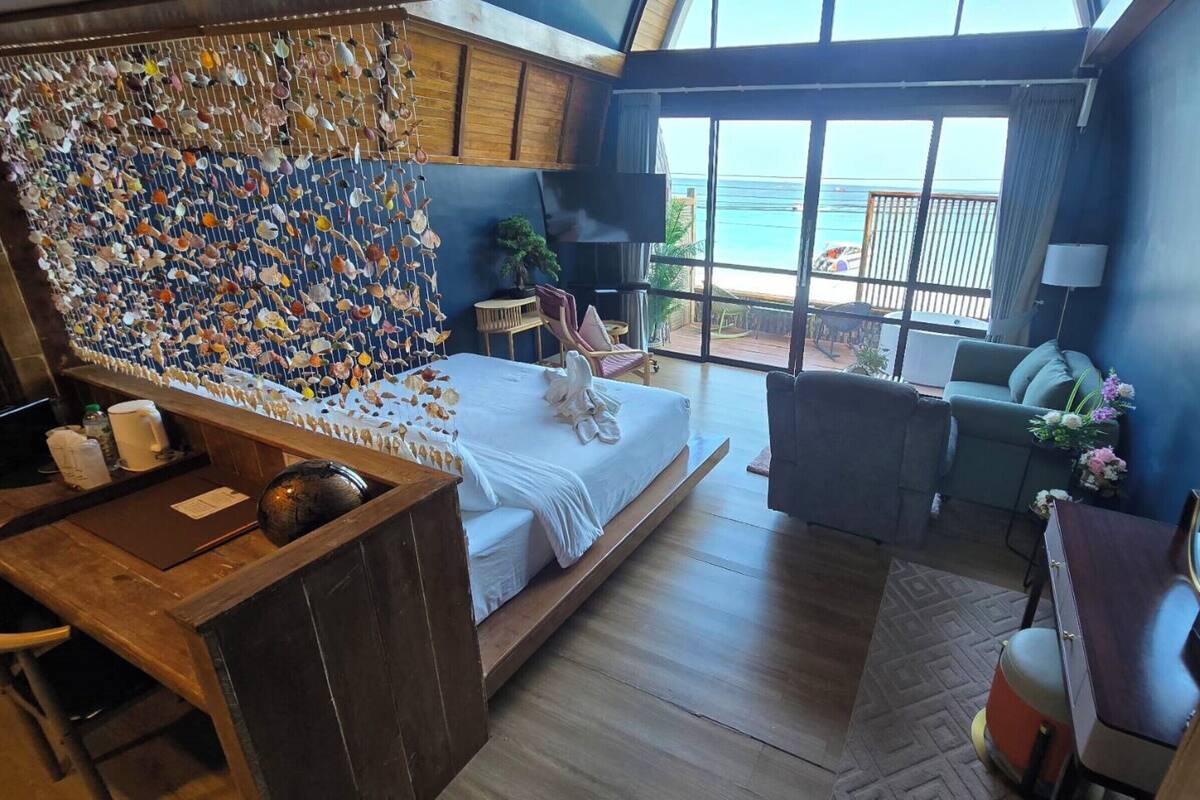Quarto com cama, escrivaninha de madeira, grande janela com vista para o oceano e parede decorada com conchas.