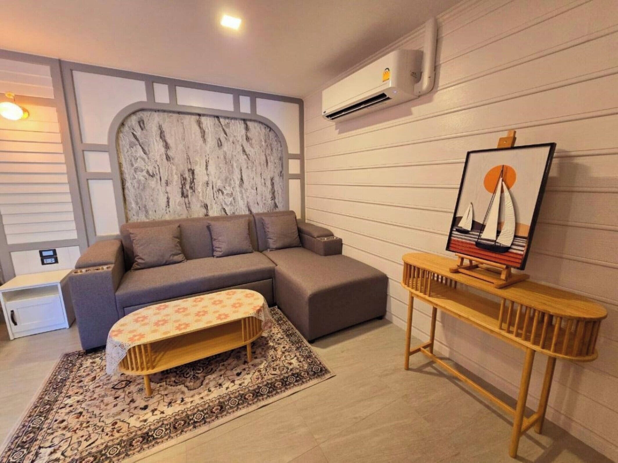 A modern living room with a grey sectional sofa, a small wooden table with a sailboat painting, and a patterned rug.