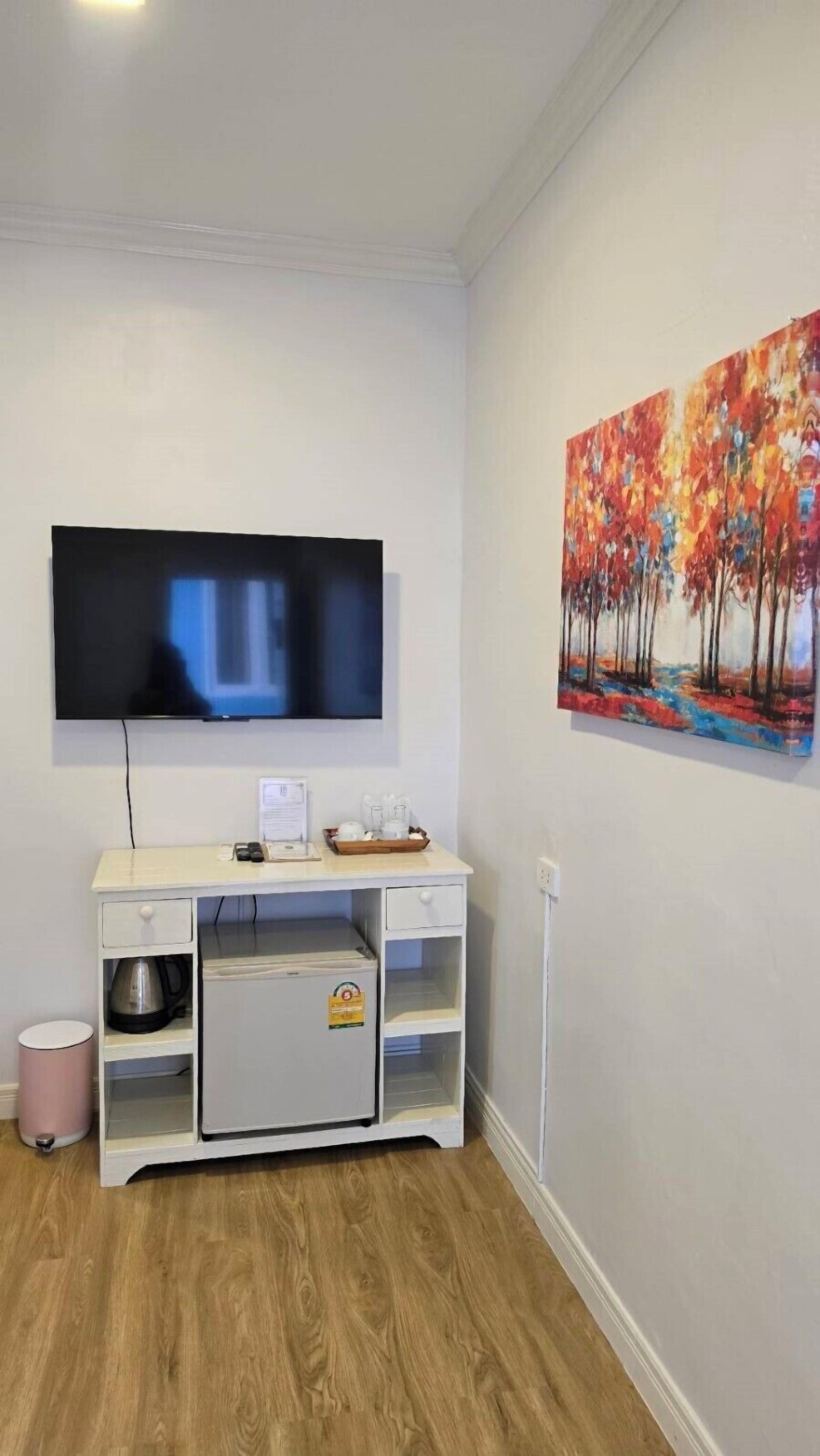 Ein Zimmer mit einem an der Wand befestigten, weißen Fernseher, einem weißen Schrank mit Regalen, einem rosa Mülleimer und einem Baumgemälde.