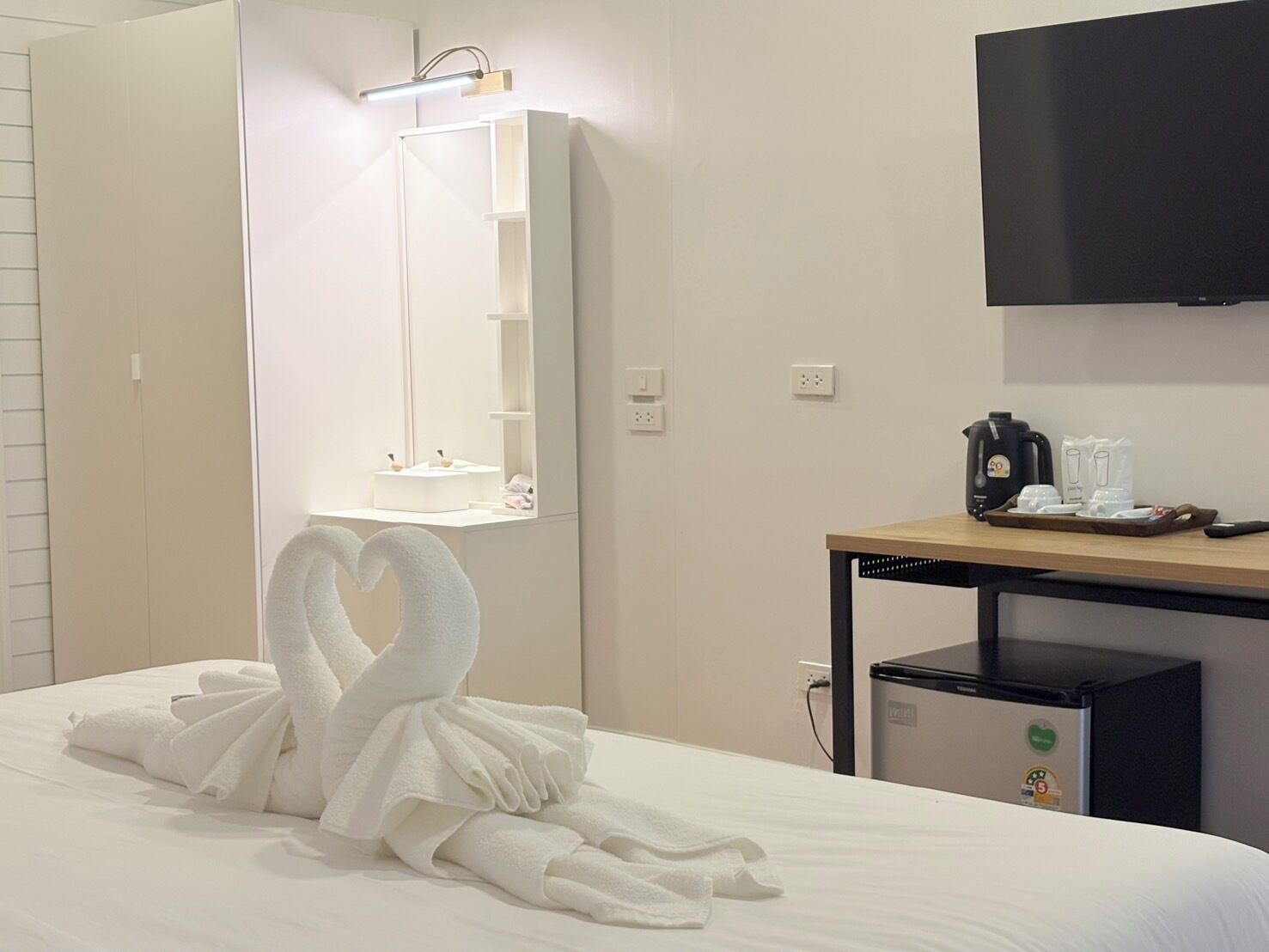 A hotel room with a bed, a bedside table with a coffee maker, a TV, and a mirror with a light above it.