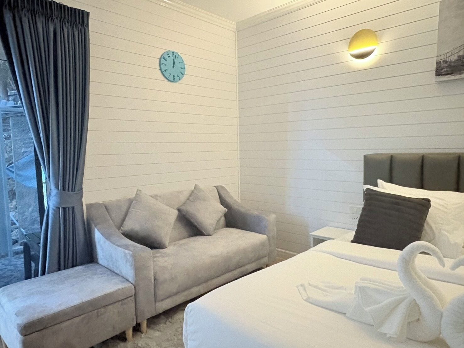 A hotel room with a bed, a sofa, a clock, and a wall-mounted light fixture.